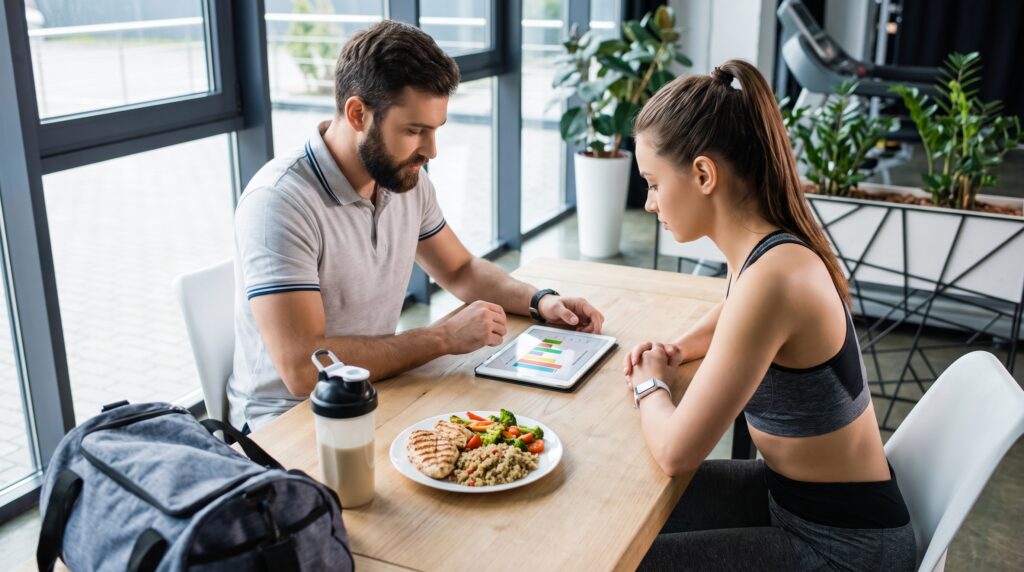 Un coach sportif et son client attablés devant une tablette, un repas équilibré et un shaker, en train de revoir un accompagnement nutritionnel